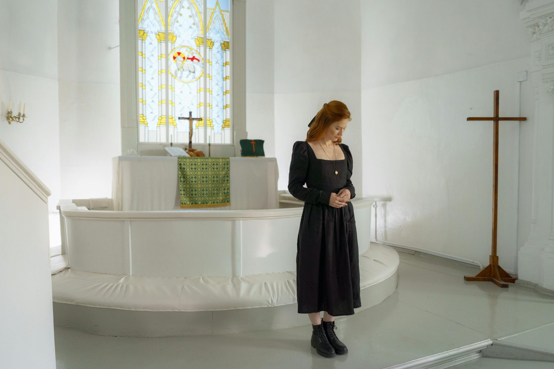 Woman in black dress standing in a church near an altar with stained glass window.