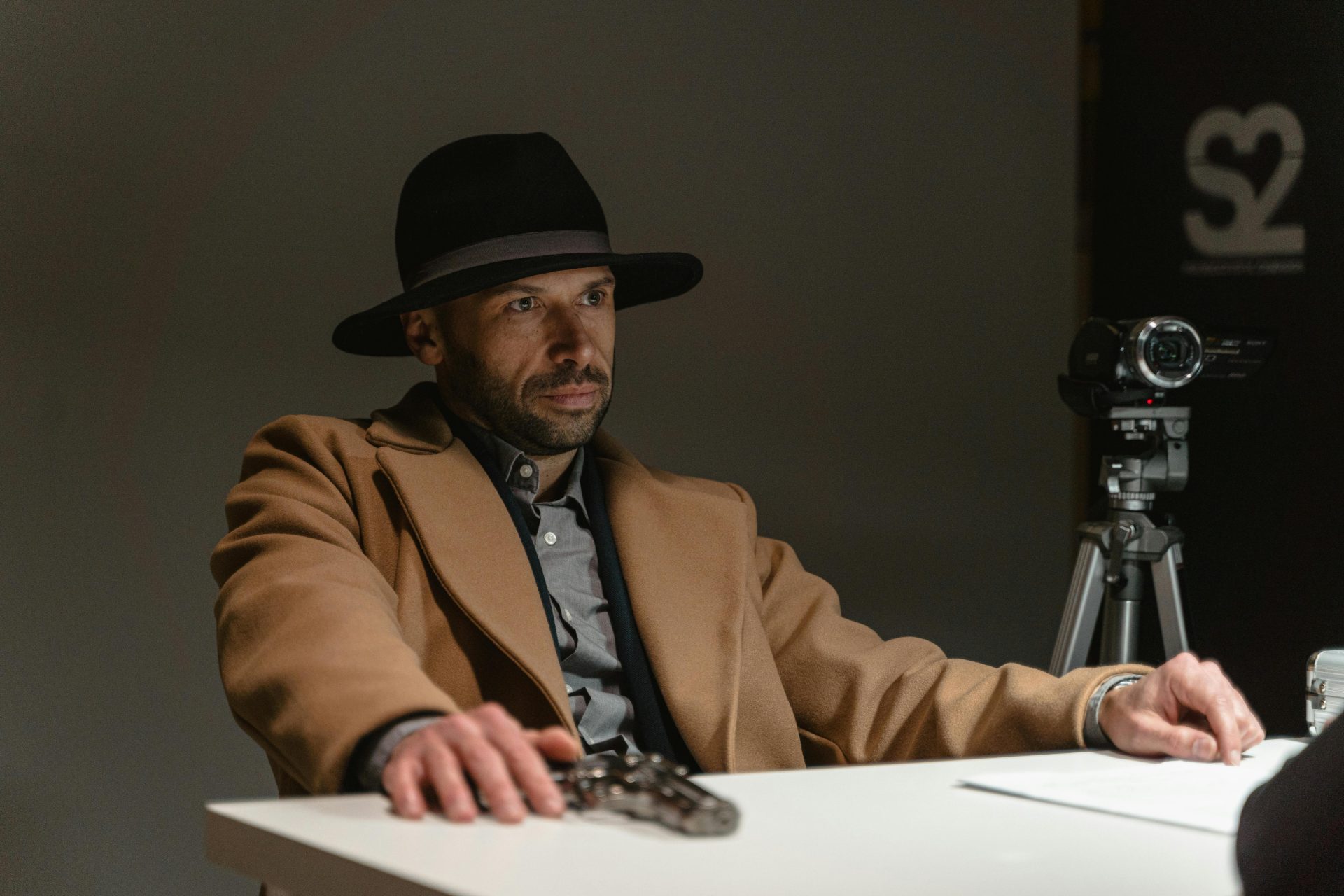 Man in a hat and coat sitting by a table with recording equipment, mysterious ambiance.
